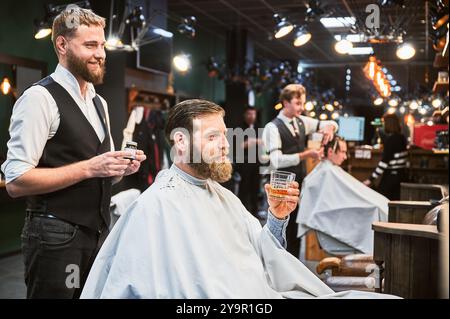 Clienti barbuti che si godono un drink mentre si fanno un nuovo taglio di capelli. Parrucchiere esperto che crea un nuovo taglio per il suo giovane cliente. Bel parrucchiere che fa capelli eleganti. Foto Stock