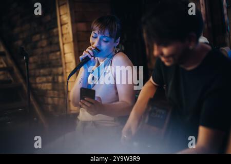 Giovane donna che canta alla serata a microfono aperto con chitarrista in un accogliente ambiente rustico da bar, creando un'atmosfera musicale vivace e intima Foto Stock