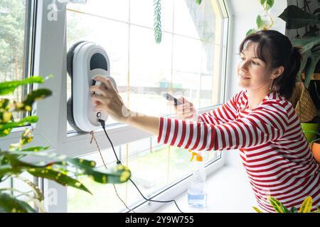 Una donna lancia un robot per la pulizia delle finestre della casa all'interno dell'interno con piante per la casa. Robotica domestica, assistente domestico, servizio di pulizia Foto Stock