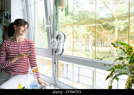 Una donna lancia un robot per la pulizia delle finestre della casa all'interno dell'interno con piante per la casa. Robotica domestica, assistente domestico, servizio di pulizia Foto Stock