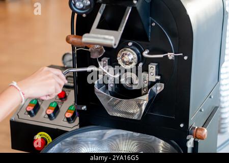 Primo piano di una macchina per la torrefazione del caffè con una mano che aziona la leva. La macchina è dotata di componenti metallici, una camera di tostatura trasparente, e. Foto Stock