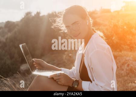 Donna laptop natura - la donna sorridente utilizza il laptop all'aperto su una collina erbosa con il sole che splende dietro di lei. Foto Stock