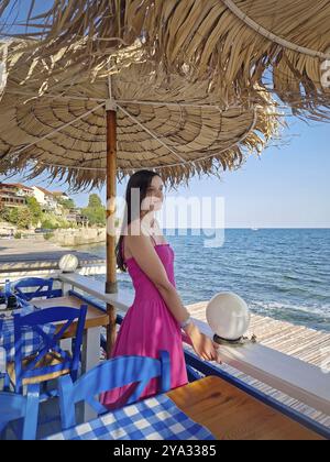 Scena serena con una graziosa giovane donna che indossa un abito rosa in piedi sotto un'ombra d'epoca su un Cafe Lodge con vista sulla costa, ammirando il b Foto Stock