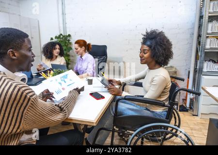 Vista laterale di una allegra donna africana con disabilità seduta su sedia a rotelle e di un collega africano maschio che lavora in un coworking Foto Stock