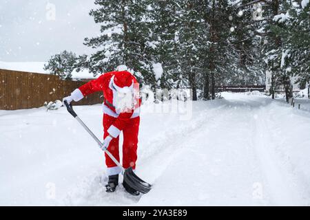 Babbo Natale pulisce la neve con la pala in inverno all'aperto dopo una nevicata. Pulire le strade del villaggio, pulire il passaggio per le auto, difficile Foto Stock