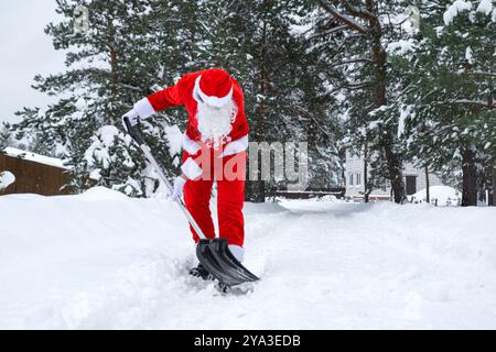 Babbo Natale pulisce la neve con la pala in inverno all'aperto dopo una nevicata. Pulire le strade del villaggio, pulire il passaggio per le auto, difficile Foto Stock