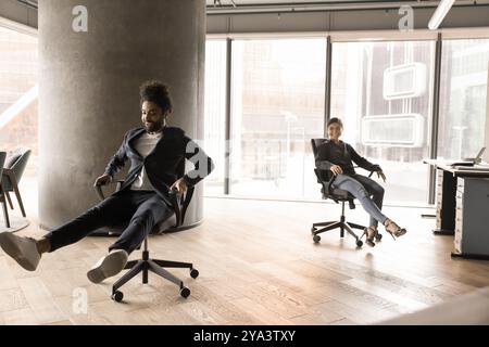 Allegri ed eccitati colleghi indiani e arabi che si divertono, cavalcando le sedie da ufficio Foto Stock