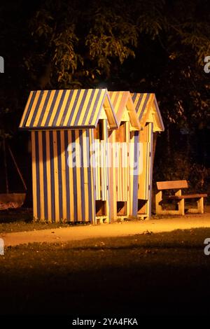 Tre cabine a strisce blu e bianche, illuminate dal semaforo in un parco notturno, con una panchina pubblica nelle vicinanze. Atmosfera calma e misteriosa. Foto Stock