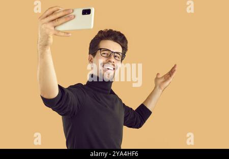 Uomo con barba felice in dolcevita nero su sfondo beige per scattare selfie su smartphone. Foto Stock