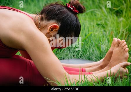 La giovane donna bianca bruna che si allunga dopo la pratica dello yoga tocca le mani con la suola dei piedi indossando un bel vestito rosso confortevole Foto Stock