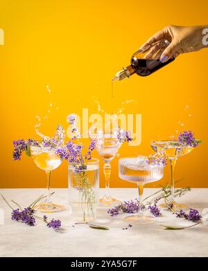 Versare a mano lo sciroppo da una bottiglia in un bicchiere da cocktail decorato con fiori di lavanda su sfondo giallo Foto Stock