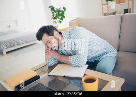 Foto di un bellissimo uomo adulto di età matura che studia a casa in un salotto interno con un enorme divano che dorme stanco durante il seminario Foto Stock