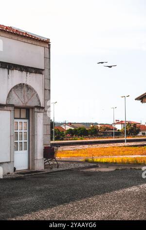 Una scena urbana illuminata dal sole mostra parte di un vecchio edificio bianco con una porta chiusa, un carrello parcheggiato e due uccelli che volano nel cielo. Binari ferroviari A. Foto Stock