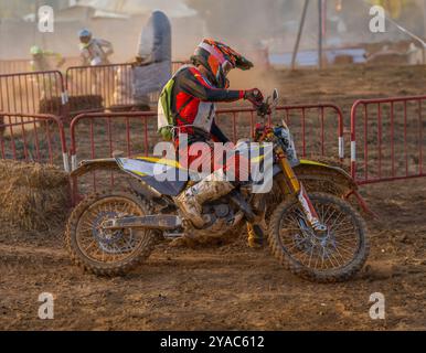 Vista laterale di un pilota di motocross in azione che prende una svolta su una pista sterrata durante una gara di moto di resistenza con altri piloti che lo inseguono Foto Stock