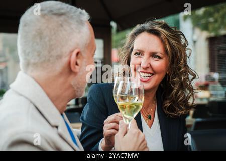 Donna matura sorridente che brinda con un bicchiere di vino bianco, godendosi un momento di gioia con il marito in un ristorante all'aperto, festeggiando e condividendo un'occasione speciale con bevande e buona compagnia. Foto di alta qualità Foto Stock