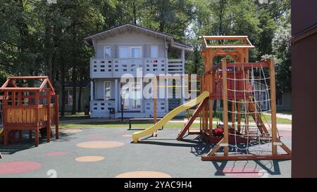 Parco giochi in legno multicolore con scivolo, bar orizzontale e cornice per arrampicate sullo sfondo di una casa bianca a due piani e alberi verdi Foto Stock