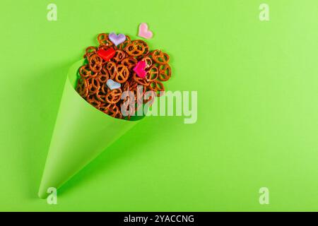 Striscione con Mini pretzel e piccoli cuori colorati in un cono di carta verde chiaro si trova su uno sfondo verde brillante con spazio per le copie. Foto Stock