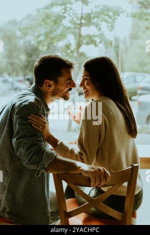 La coppia si appoggia, condividendo risate e dolci sussurri, mentre la luce del sole filtra attraverso la finestra del caffè, evidenziando la loro connessione gioiosa Foto Stock