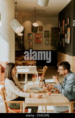 Una giovane coppia condivide un momento di cuore in una caffetteria, sorridendo e tenendo per mano le bevande mentre è circondata da un'atmosfera affascinante e artistica. Foto Stock