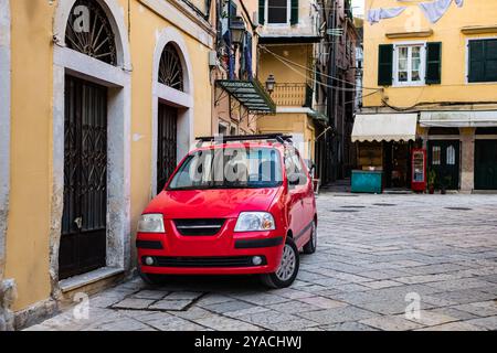 Stradina stretta della città vecchia con case colorate e auto rossa parcheggiata su un lato della città europea di Corfù in Grecia. Una piccola auto rossa per strada con un vecchio amico Foto Stock