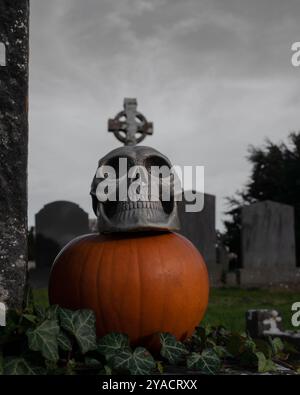 Teschio umano spaventoso con una croce celtica in cima alla testa con una zucca a terra con un cupo cimitero sullo sfondo, il 31 ottobre Foto Stock