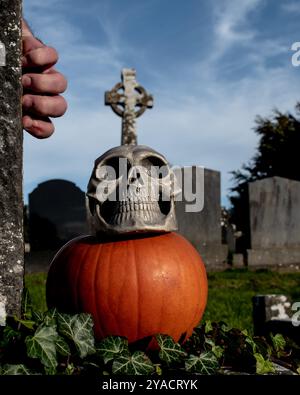 Spaventoso teschio umano di halloween con una croce celtica sulla testa con una zucca a terra e una mano spaventosa con un dito sanguinante nel cimitero Foto Stock