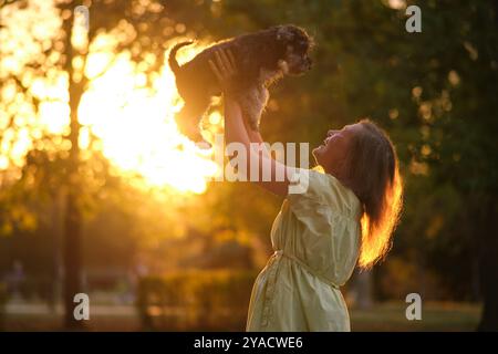 Donna felice che solleva un piccolo cane nel parco durante il tramonto. Fotografia di ritratti all'aperto con retroilluminazione naturale. Momenti di gioia con animali domestici e natura convivono Foto Stock