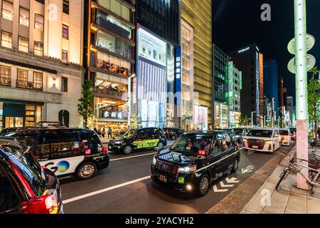 Vista serale lungo la via Ginza con molti taxi che passano davanti a vari alti negozi. Lo store nero Hublot sullo sfondo. Foto Stock