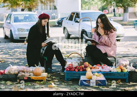 Romny, Ucraina, 29 settembre 2019: Giovani donne che leggono un libro e vendono verdure mentre sono sedute all'aperto in un tranquillo ambiente urbano. Foto Stock