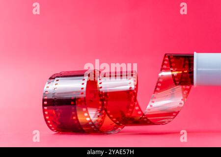 Primo piano di una bobina con una striscia di pellicola rossa che si srotola su uno sfondo rosa. La pellicola presenta perforazioni lungo i bordi, showcasi Foto Stock