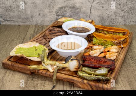 Piatto di legno ricoperto di carne asada grigliata, tacos, gamberetti, salsiccia chorizo, salsa, e cipolla verde per una festa di cibo messicano. Foto Stock