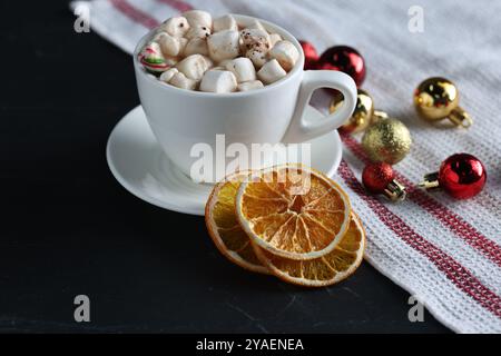 Gustosa bevanda a base di cioccolata calda con marshmallow in tazza, fette di arancia secca e baule sul tavolo nero Foto Stock