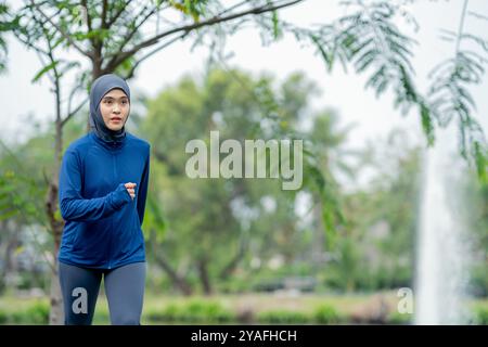 Donna musulmana asiatica che indossa abbigliamento sportivo e un hijab blu che si esercita e corre all'aperto al parco la mattina. con una sensazione di freschezza. Foto Stock