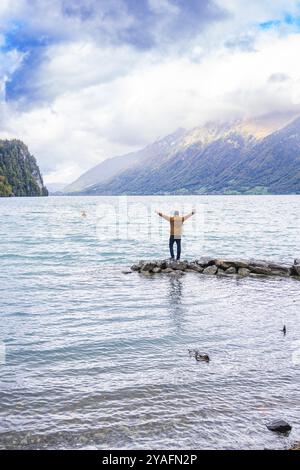 Persona in piedi con le braccia allungate sulle rocce vicino al lago con sfondo di montagna sotto il cielo spettacolare, lago di Brienz, Svizzera, Europa Foto Stock