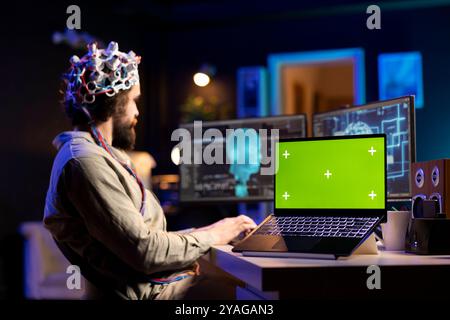 Concentratevi su un notebook con schermo verde con un uomo in background sfocato che utilizza le cuffie EEG per trasferire la mente nel mondo virtuale. Primo piano di un dispositivo di mockup accanto allo scienziato che utilizza la tecnologia per ottenere superintelligenza Foto Stock