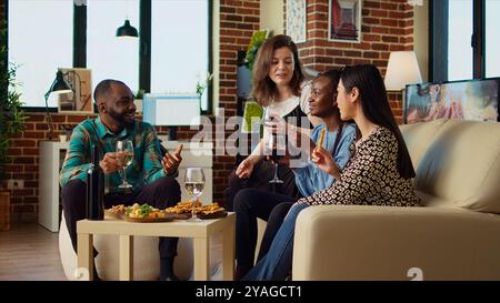 Amici che ridono, bevono vino rosso e bianco, parlano della loro vita in un accogliente soggiorno dell'appartamento. I colleghi trascorrono del tempo insieme, mangiano da salumi a casa, ingrandiscono le foto Foto Stock