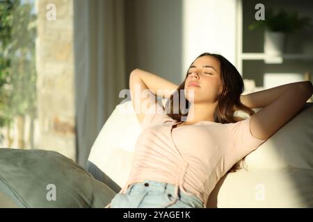 Donna rilassata seduta su un divano con le braccia sulla testa accanto a una finestra di casa Foto Stock