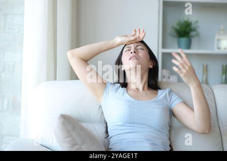 Donna asiatica stressata che soffre di colpo di calore che sventola con la mano seduta su un divano a casa Foto Stock