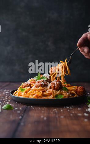 Mano con la forchetta nel piatto di spaghetti e polpette sul tavolo. Foto Stock