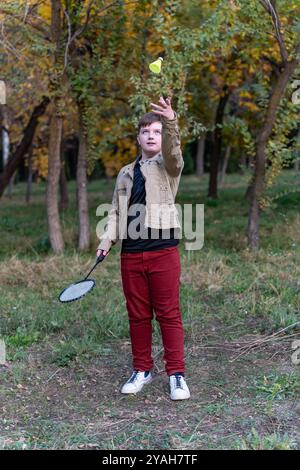 Un adolescente con giacca beige, jeans Bordeaux e sneakers bianche si trova in un parco autunnale, con una racchetta da badminton in mano destra. Foto Stock