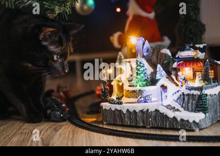 Gatto nero osserva curiosamente un villaggio natalizio in miniatura sotto l'albero. Decorazioni natalizie con ambientazione invernale illuminata e set di treni. Foto Stock