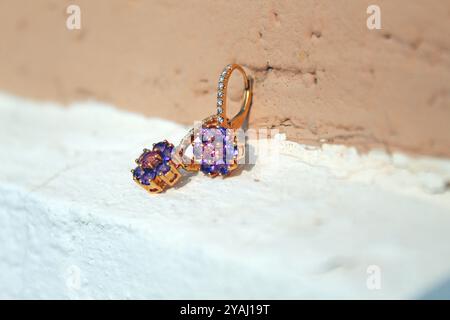 Coppia di orecchini floreali dorati adornati con piccole pietre preziose viola e rosa, appoggiati su una superficie ruvida con un morbido sfondo marrone Foto Stock