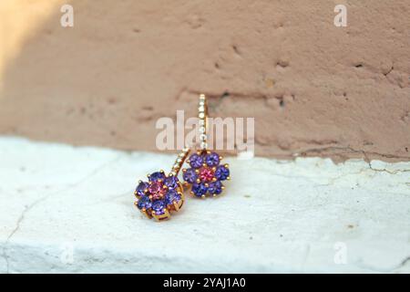 Coppia di orecchini floreali dorati adornati con piccole pietre preziose viola e rosa, appoggiati su una superficie ruvida con un morbido sfondo marrone Foto Stock