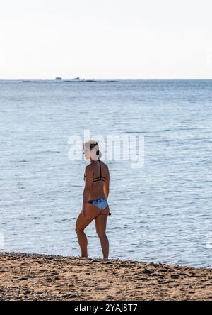 Attraente giovane donna sottile che cammina da sola lungo una spiaggia greca mediterranea con indosso un bikini blu e che guarda alla sua sinistra. Foto Stock