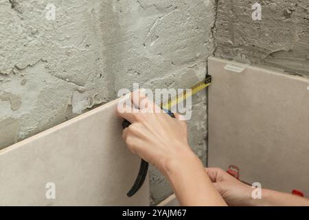 Donna che piastrella le pareti del bagno, utilizzando un metro a nastro per misurare la lunghezza. Processo di installazione delle piastrelle nel bagno passo dopo passo. Miglioramento fai da te Foto Stock