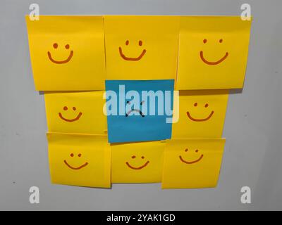 Contrasto emotivo. Nota adesiva con volto blu con espressione arrabbiata circondata da facce sorridenti su fogli gialli, su sfondo bianco. Foto Stock