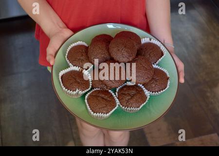 Una pila di muffin al cioccolato fatti in casa su un piatto verde tenuto dalle mani di una bambina in un vestito rosso Foto Stock