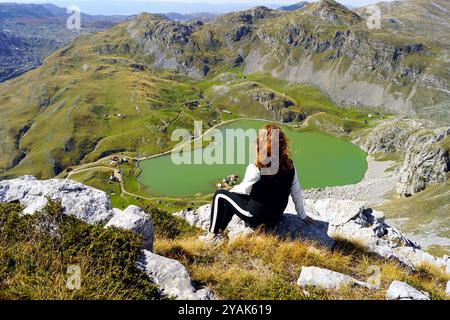 Una ragazza riposa dopo aver scalato la cima di una montagna e gode della vista. Una turista femminile siede su una roccia con il lago Kapetanovo sullo sfondo. Foto Stock