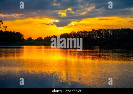 Il sole tramonta dietro l'orizzonte, proiettando vivaci sfumature arancioni e gialle sulle calme acque del lago Foto Stock