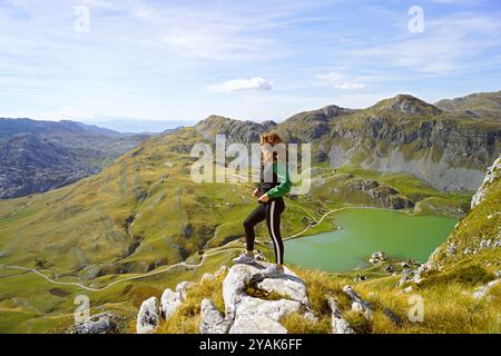 Escursionista in Montenegro: Una ragazza sta su una roccia e guarda verso una strada di montagna. Una turista femminile sullo sfondo delle montagne e del lago Kapetanovo Foto Stock